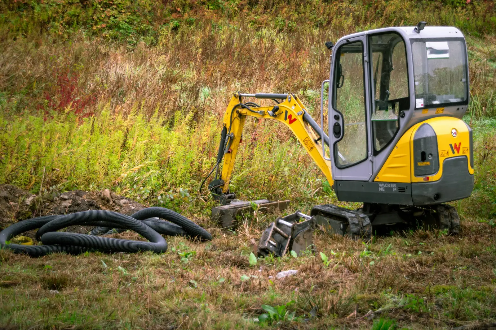 A mini digger beside coils of drainage pipe in long grass