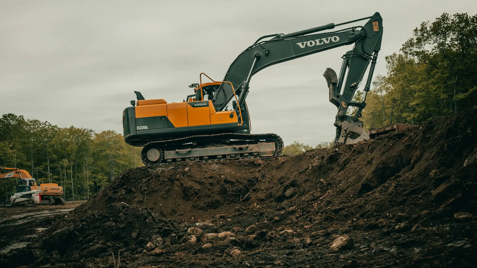 A tracked excavator working on top of a freshly dug earth bank, with trees behind
