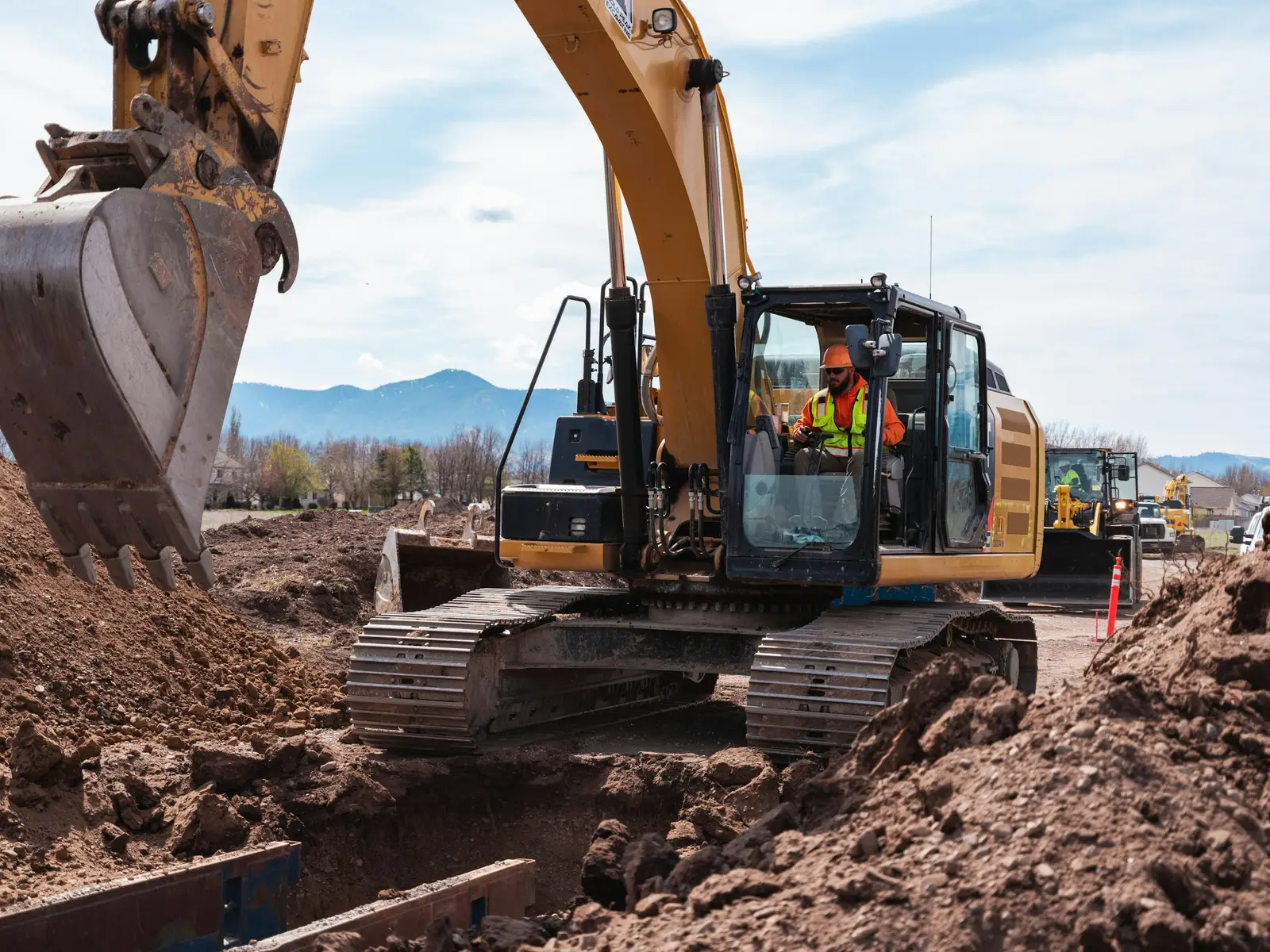 An operator in hi-vis at the controls of an excavator, beside a trench